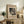May include: Framed abstract art print featuring geometric shapes, circles, and lines in black and white displayed on a rustic wooden sideboard. The beige matted poster shows Wassily Kandinsky's name at bottom. Room setting includes fern plant, ceramic vase, stacked books, throw pillows on cream chair, and warm neutral tones throughout the cozy living space.
