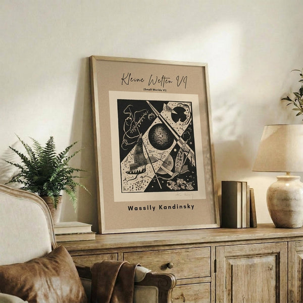 May include: Framed abstract art print featuring geometric shapes, circles, and lines in black and white displayed on a rustic wooden sideboard. The beige matted poster shows Wassily Kandinsky's name at bottom. Room setting includes fern plant, ceramic vase, stacked books, throw pillows on cream chair, and warm neutral tones throughout the cozy living space.