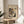May include: Framed Kandinsky abstract art print featuring colorful geometric shapes, lines and forms in an oval composition against a dark background. The cream-colored poster is displayed in a thin wooden frame on a light wood round side table next to a beige upholstered chair. Natural sunlight filters through white curtains, casting soft shadows across the neutral-toned room setting with a plant visible in the corner.
