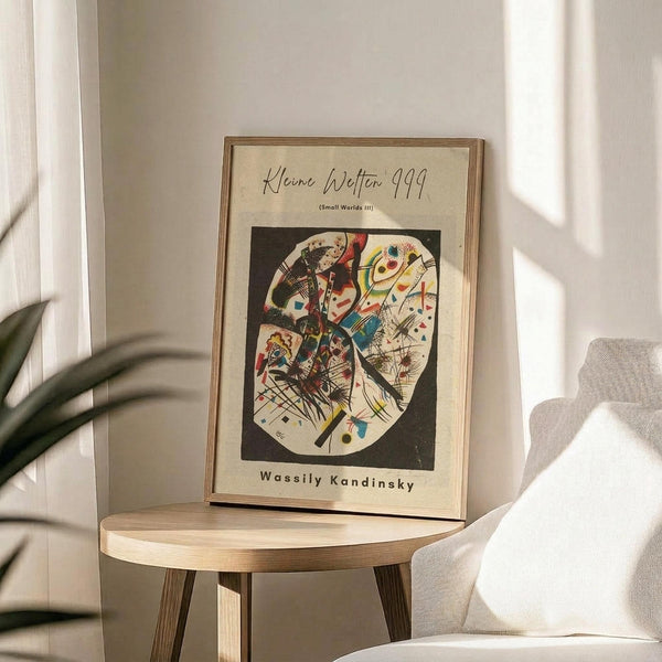 May include: Framed Kandinsky abstract art print featuring colorful geometric shapes, lines and forms in an oval composition against a dark background. The cream-colored poster is displayed in a thin wooden frame on a light wood round side table next to a beige upholstered chair. Natural sunlight filters through white curtains, casting soft shadows across the neutral-toned room setting with a plant visible in the corner.