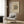 May include: Abstract geometric artwork poster with black linear designs and circular forms on cream background, displayed above beige upholstered chair with geometric patterned pillow. Modern brass floor lamp with white circular base stands nearby next to dark round side table with open book. Warm wood-paneled wall backdrop with natural lighting from window with white curtains.