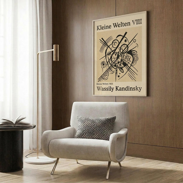 May include: Abstract geometric artwork poster with black linear designs and circular forms on cream background, displayed above beige upholstered chair with geometric patterned pillow. Modern brass floor lamp with white circular base stands nearby next to dark round side table with open book. Warm wood-paneled wall backdrop with natural lighting from window with white curtains.
