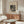 May include: Abstract black and white geometric artwork by Wassily Kandinsky displayed above a wooden sideboard in a minimalist living room. The framed print features circular and angular shapes in a modernist style. Room shows cream-colored chair, glass oval coffee table, white mushroom lamp, and copper floor lamp against neutral walls.