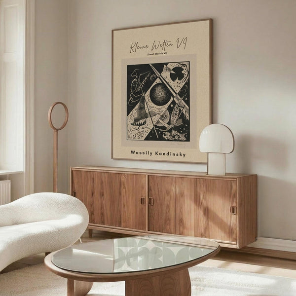 May include: Abstract black and white geometric artwork by Wassily Kandinsky displayed above a wooden sideboard in a minimalist living room. The framed print features circular and angular shapes in a modernist style. Room shows cream-colored chair, glass oval coffee table, white mushroom lamp, and copper floor lamp against neutral walls.
