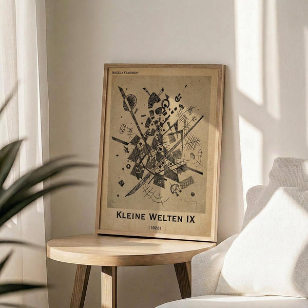 May include: Framed abstract artwork by Wassily Kandinsky displayed on a round wooden side table next to a beige upholstered chair. The print features black geometric shapes, lines, and patterns on aged beige paper in a light wood frame. Soft natural lighting creates shadows on white walls, with green plant leaves visible in the corner of the minimalist interior setting.