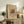 May include: Framed abstract art print with colorful geometric shapes and lines on beige background, displayed on rustic wooden sideboard next to fern plant, ceramic vase, and stacked books in warm-toned living room setting with cream upholstered chair visible in foreground.