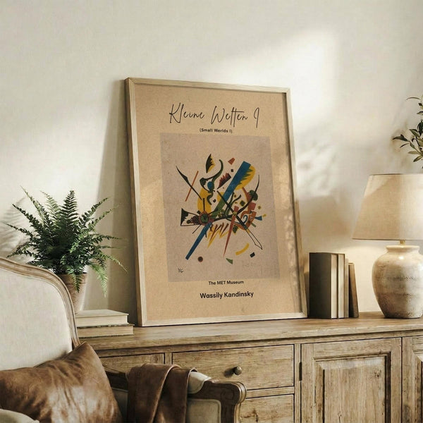 May include: Framed abstract art print with colorful geometric shapes and lines on beige background, displayed on rustic wooden sideboard next to fern plant, ceramic vase, and stacked books in warm-toned living room setting with cream upholstered chair visible in foreground.