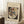 May include: Framed art print leaning against white wall corner on fluffy white rug. Shows abstract geometric composition with colorful shapes, lines and forms in black border on cream background. Wassily Kandinsky attribution text below artwork. Wooden side table with white coffee cup and saucer visible on left. Natural lighting creates soft shadows on wall.