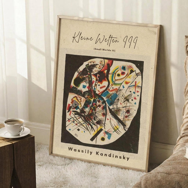 May include: Framed art print leaning against white wall corner on fluffy white rug. Shows abstract geometric composition with colorful shapes, lines and forms in black border on cream background. Wassily Kandinsky attribution text below artwork. Wooden side table with white coffee cup and saucer visible on left. Natural lighting creates soft shadows on wall.