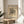 May include: Framed abstract art print with black geometric lines, curves and organic shapes on cream background, displayed on round wooden side table next to beige upholstered chair. Natural lighting creates soft shadows on white wall behind, with plant leaves visible in corner of minimalist interior setting.
