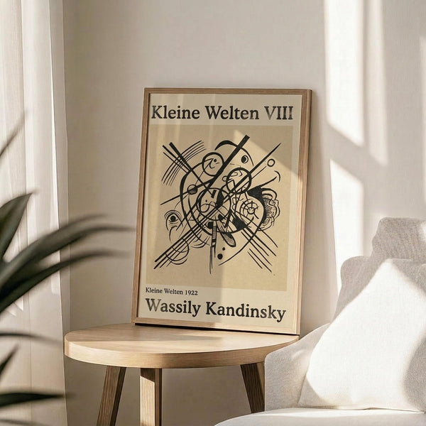 May include: Framed abstract art print with black geometric lines, curves and organic shapes on cream background, displayed on round wooden side table next to beige upholstered chair. Natural lighting creates soft shadows on white wall behind, with plant leaves visible in corner of minimalist interior setting.