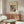 May include: Abstract art poster by Wassily Kandinsky displayed above a wooden sideboard in a modern living room. The vintage-style print features colorful geometric shapes and lines within a circular composition on cream paper with dark border. White curved chair and glass oval coffee table are positioned nearby, with warm natural lighting creating shadows across the minimalist space.