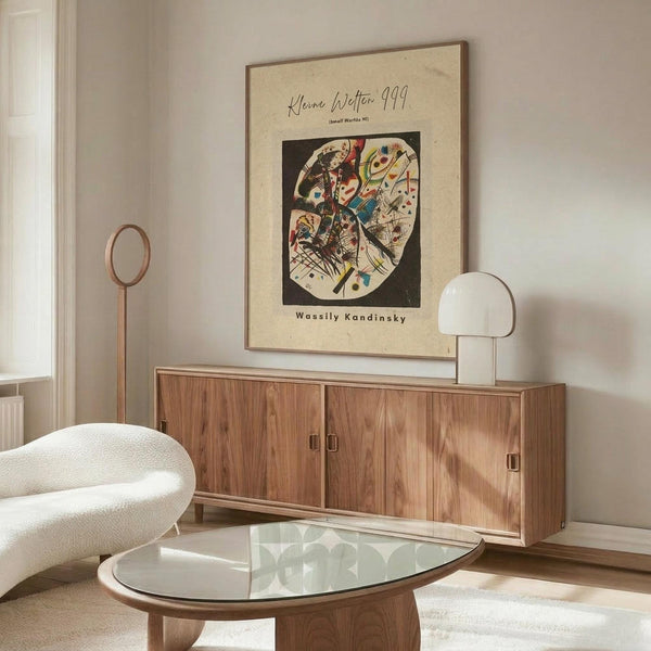 May include: Abstract art poster by Wassily Kandinsky displayed above a wooden sideboard in a modern living room. The vintage-style print features colorful geometric shapes and lines within a circular composition on cream paper with dark border. White curved chair and glass oval coffee table are positioned nearby, with warm natural lighting creating shadows across the minimalist space.