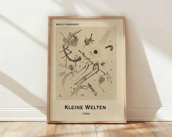 May include: Framed abstract art print featuring geometric shapes, lines, and symbols in black ink on cream-colored paper, displayed against white wall on light wooden floor. The artwork shows Kandinsky's distinctive style with triangles, circles, dots, curved lines, and crosshatched patterns scattered across the composition. Natural wood frame with warm lighting casting soft shadows.