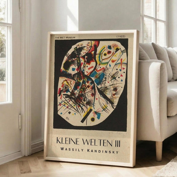 May include: Framed abstract art poster leaning against white wall next to beige sofa in bright room with large windows. The artwork features Kandinsky's colorful geometric composition with red, blue, yellow, and black shapes on cream background. Text reads "THE MET MUSEUM (1922)" and "KLEINE WELTEN III WASSILY KANDINSKY" below the oval-shaped abstract design. Light wood frame, hardwood floors visible.