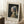 May include: Framed reproduction of Vermeer's Girl with a Pearl Earring displayed on white fluffy rug against white wall with window light. Print shows young woman in blue and yellow headwrap with pearl earring on dark background. Wooden frame with cream matting, positioned next to small wooden side table with white coffee cup and saucer.