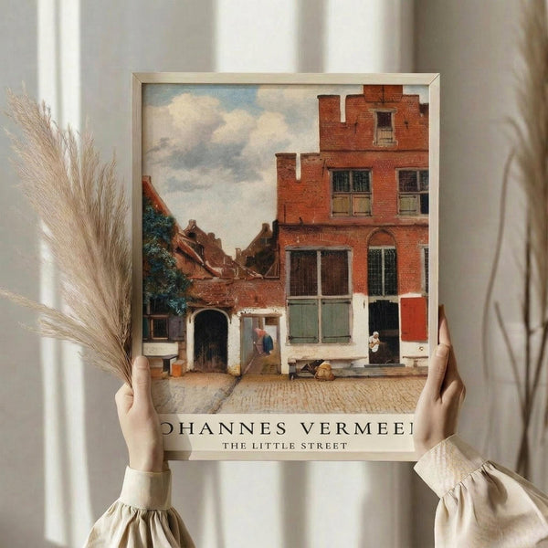 May include: Person in cream clothing holding framed art print of Dutch street scene with red brick buildings, cobblestone courtyard, and cloudy sky. Print shows classic 17th century architecture with shuttered windows and small figures. Dried wheat stems visible on left side against soft neutral background with natural window lighting.