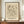 May include: Framed abstract art print displayed on wooden credenza beside white ceramic vase and stacked books. The beige-toned artwork features geometric lines, shapes, and scattered elements in black ink drawing style. Simple brown frame against cream wall with white molding. Print shows artist name at top and German title with date at bottom in clean typography.