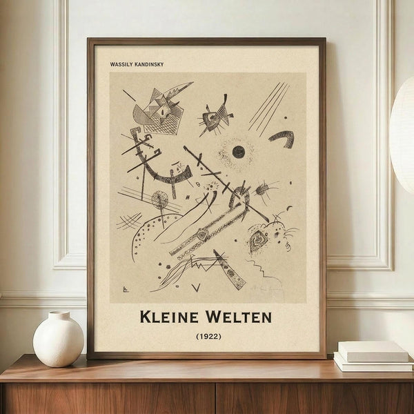 May include: Framed abstract art print displayed on wooden credenza beside white ceramic vase and stacked books. The beige-toned artwork features geometric lines, shapes, and scattered elements in black ink drawing style. Simple brown frame against cream wall with white molding. Print shows artist name at top and German title with date at bottom in clean typography.
