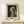 May include: Framed art print of Vermeer's Girl with a Pearl Earring displayed on light wood surface against cream wall. The classic portrait shows a young woman in blue and yellow headwrap with pearl earring, looking over her shoulder against dark background. Print has cream matting in natural wood frame, with small white gift box tied with greenery positioned in front.