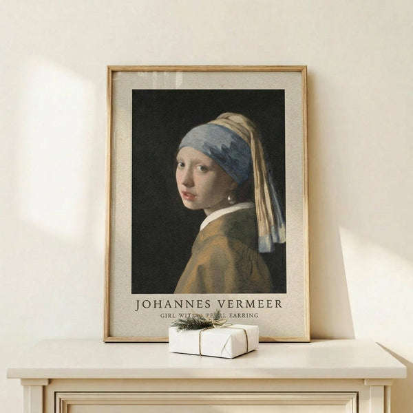 May include: Framed art print of Vermeer's Girl with a Pearl Earring displayed on light wood surface against cream wall. The classic portrait shows a young woman in blue and yellow headwrap with pearl earring, looking over her shoulder against dark background. Print has cream matting in natural wood frame, with small white gift box tied with greenery positioned in front.