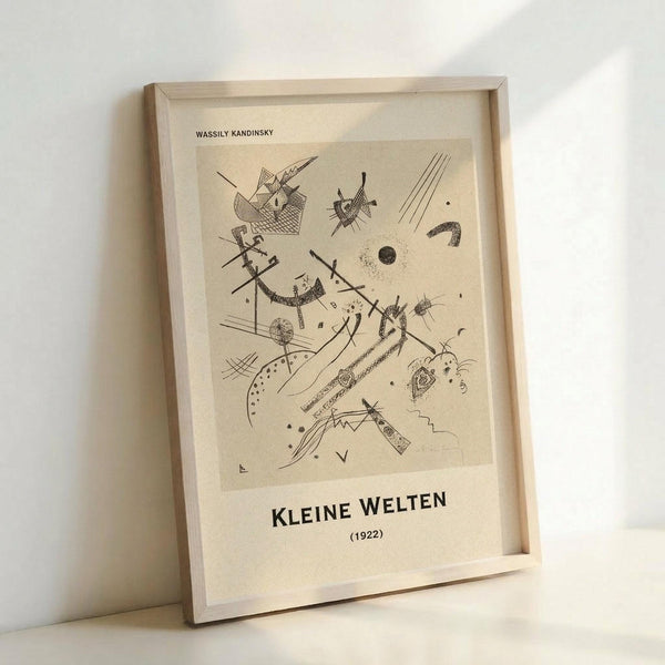 May include: Framed abstract art print in light wood frame leaning against white wall. Black line drawing on cream background features geometric shapes, curved lines, dots, and angular forms in Kandinsky's distinctive abstract style. Text reads "Wassily Kandinsky" at top and "Kleine Welten (1922)" at bottom. Minimalist presentation shows fine art reproduction with clean matting.