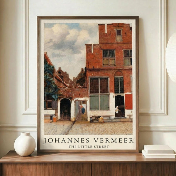 May include: Framed art print of a Dutch Golden Age street scene with red brick buildings, cobblestone courtyard, and small figures in period clothing under a cloudy sky. The poster is displayed on a wooden sideboard next to a white ceramic vase and stacked books in a neutral-toned room with white paneled walls.