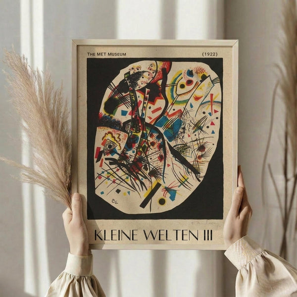 May include: Hands holding a framed abstract art print featuring vibrant geometric shapes, lines, and colors in red, blue, yellow, and black on cream background. Light wood frame with museum text header and "KLEINE WELTEN III" at bottom. Person wearing cream ruffle-sleeve blouse holds dried pampas grass, standing by white curtained window in soft natural light.