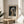 May include: Framed art print of Vermeer's Girl with a Pearl Earring displayed on a round wooden side table next to a beige upholstered chair. The print shows a young woman in blue and yellow headwrap with pearl earring against dark background, housed in thin gold frame with white matting. Soft natural light creates shadows across neutral-toned room with plant leaves visible in corner.