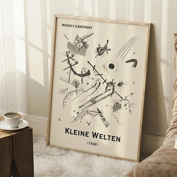 May include: Framed abstract art print in light wood frame leaning against white wall corner. Black line drawings on cream background show geometric shapes, arrows, circles, and angular forms in Kandinsky's distinctive style. Text reads "Wassily Kandinsky" and "Kleine Welten (1922)". Coffee cup on wooden block and beige blanket visible nearby on white textured rug.