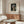 May include: Framed art print of Vermeer's Girl with a Pearl Earring displayed above a warm wood sideboard in a minimalist living room. The reproduction shows a young woman in blue and yellow headwrap with pearl earring against dark background. Modern furniture includes white curved chair, glass oval coffee table, and brass floor lamp. Neutral beige walls with natural lighting.