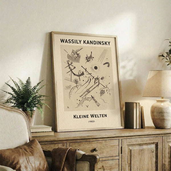 May include: Framed Kandinsky abstract art print in light wood frame, leaning against white wall on rustic wooden sideboard. Black line drawing features geometric shapes, curves, and abstract forms on cream background. Styled with fern plant, ceramic vase, stacked books, and beige upholstered chair with brown throw pillow in warm, minimalist living room setting.