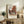 May include: Framed art print of Dutch street scene with red brick buildings and cobblestone courtyard displayed on rustic wooden sideboard. Warm neutral room setting with fern plant, ceramic vase, stacked books, and beige upholstered chair. Print shows classic European architecture with multi-story building featuring traditional windows and doorways under cloudy sky.