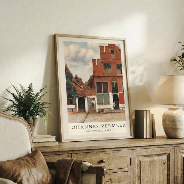 May include: Framed art print of Dutch street scene with red brick buildings and cobblestone courtyard displayed on rustic wooden sideboard. Warm neutral room setting with fern plant, ceramic vase, stacked books, and beige upholstered chair. Print shows classic European architecture with multi-story building featuring traditional windows and doorways under cloudy sky.
