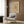 May include: Framed beige poster with abstract black line drawings and geometric shapes hanging on wood-paneled wall above cream upholstered armchair with geometric patterned pillow. Modern floor lamp with brass finish and round base stands nearby next to dark round side table with open book. Neutral toned room with natural lighting from window with curtains.