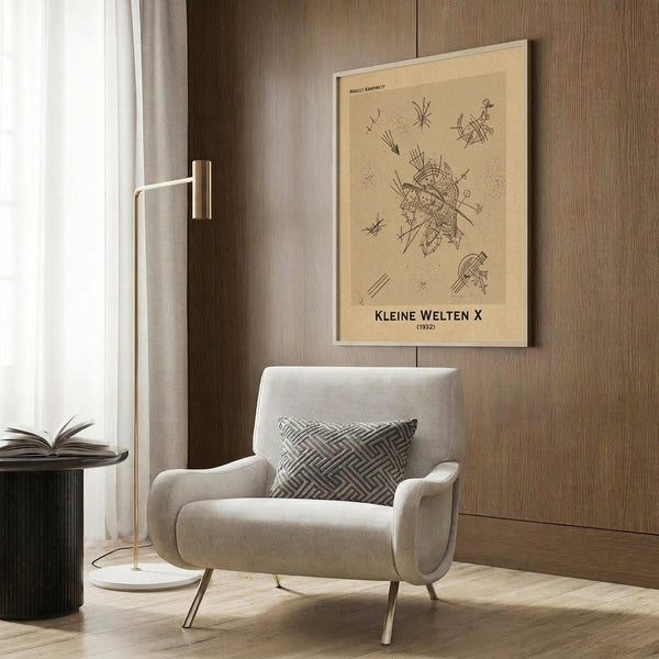 May include: Framed beige poster with abstract black line drawings and geometric shapes hanging on wood-paneled wall above cream upholstered armchair with geometric patterned pillow. Modern floor lamp with brass finish and round base stands nearby next to dark round side table with open book. Neutral toned room with natural lighting from window with curtains.