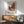 May include: Framed Vermeer reproduction showing Dutch street scene with red brick buildings and cloudy sky, displayed above wooden sideboard with white ceramic lamp in modern living room with sage green mid-century chair and natural lighting casting shadows on white wall.