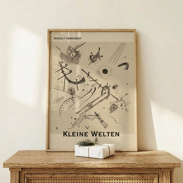 May include: Framed abstract art print in light wood frame leaning against white wall on rustic wooden surface. Black line drawing on beige background features geometric shapes, curved lines, dots, and angular forms in Kandinsky's distinctive abstract style. Small white wrapped gift with twine and greenery sits beside frame. Warm, minimalist home decor setting with natural wood textures.