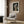 May include: Framed art print of Vermeer's Girl with a Pearl Earring hanging on wood-paneled wall above beige upholstered armchair with geometric patterned pillow. Modern brass floor lamp stands beside black round side table with open book. Neutral toned room with natural lighting from window with sheer curtains.