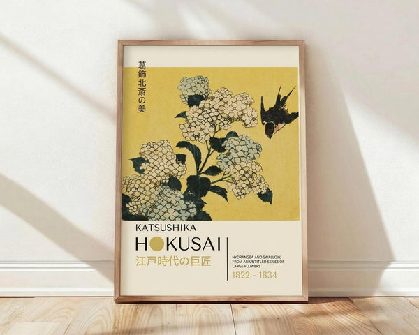 May include: Framed Japanese art print leaning against white wall on light wood floor. Yellow-toned artwork shows detailed hydrangea flowers with dark green leaves and a black swallow bird in flight. Modern wooden frame surrounds the vintage botanical illustration. Soft natural lighting creates diagonal shadows across the scene.