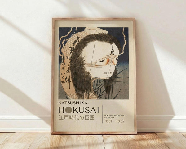May include: Framed poster print featuring a Japanese woodblock art style illustration of a ghostly female figure with long black hair, pale skin, and traditional clothing. The artwork shows dramatic lighting and classic ukiyo-e aesthetics with Japanese text at the bottom. The print is displayed in a light wooden frame, leaning against a white wall on light wooden flooring with soft natural lighting casting shadows across the scene.