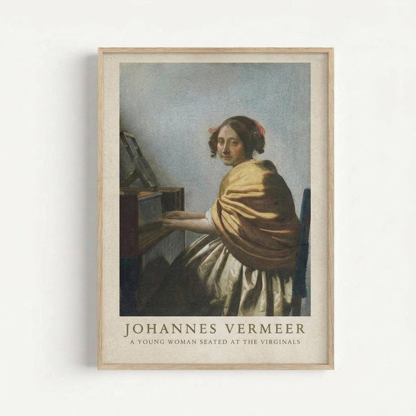 Framed art print of a classical painting showing a young woman in golden and white robes seated at a wooden desk with an open book or music stand. The woman has dark hair with red ribbons and is positioned in three-quarter view against a muted blue-gray background. Below the image is text reading "JOHANNES VERMEER" and "A YOUNG WOMAN SEATED AT THE VIRGINALS" in serif font on cream-colored matting. The print is displayed in a light wooden frame against a white wall.