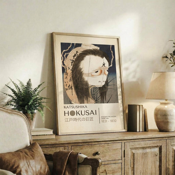 May include: Framed Japanese woodblock print featuring a ghostly female figure with long black hair and pale white face, displayed on a rustic wooden credenza alongside fern plant, books, and ceramic vase in warm-toned living room setting with beige upholstered furniture