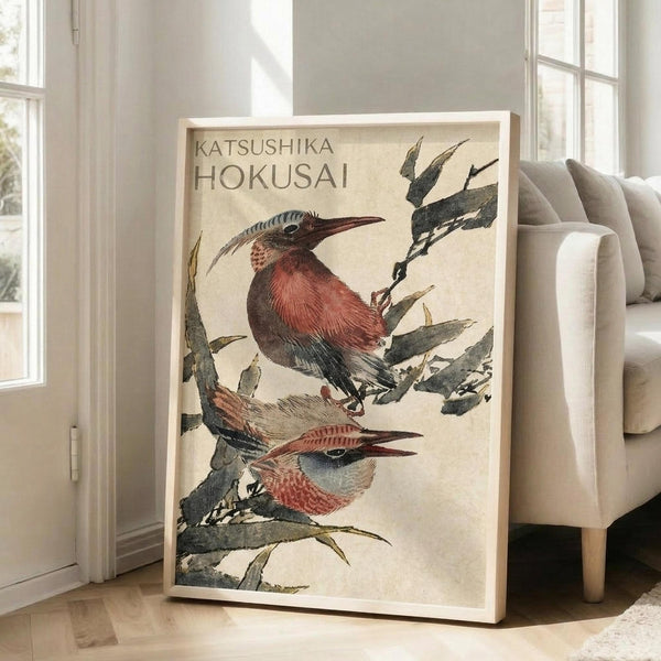 May include: Framed art print featuring two colorful birds perched on leafy branches in traditional Japanese woodblock style, displayed against white wall next to beige upholstered chair in bright living room with large windows and hardwood floors. The birds show rich reddish-brown plumage with detailed feather textures on cream background.