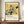 May include: Framed museum-style poster featuring Japanese woodblock print of white hydrangea flowers with dark green leaves and black swallow on golden yellow background. Vertical Japanese text and "KATSUSHIKA HOKUSAI" with dates 1822-1834 below artwork. Brown wooden frame displayed on walnut sideboard beside white ceramic vase and stacked books in bright modern room with white paneled walls.