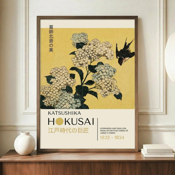 May include: Framed museum-style poster featuring Japanese woodblock print of white hydrangea flowers with dark green leaves and black swallow on golden yellow background. Vertical Japanese text and "KATSUSHIKA HOKUSAI" with dates 1822-1834 below artwork. Brown wooden frame displayed on walnut sideboard beside white ceramic vase and stacked books in bright modern room with white paneled walls.