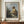 May include: Framed art print of Vermeer's classical painting showing a young woman in golden yellow shawl and white dress seated at virginals, looking over shoulder. Brown wooden frame with white matting displays on warm wood sideboard beside white ceramic vase and stacked books against cream paneled wall.