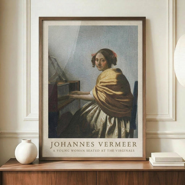 May include: Framed art print of Vermeer's classical painting showing a young woman in golden yellow shawl and white dress seated at virginals, looking over shoulder. Brown wooden frame with white matting displays on warm wood sideboard beside white ceramic vase and stacked books against cream paneled wall.