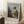 May include: Framed art print of Vermeer's classical painting showing a young woman in yellow and white robes seated at a virginals keyboard instrument, displayed on white textured rug against bright white wall with window light, alongside wooden side table with coffee cup and beige throw blanket.
