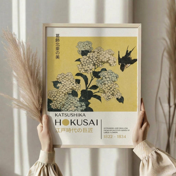 May include: Person in beige clothing holding framed Japanese woodblock print featuring white hydrangea flowers and black swallow on yellow background. Print shows museum-style typography with artist name Katsushika Hokusai and dates 1822-1834. Light wood frame against white curtained window with dried pampas grass visible on left side.